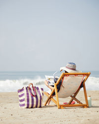 A woman sits on the beach with the Beach Tote with Flat Clutch in Grape and Shell Stripe