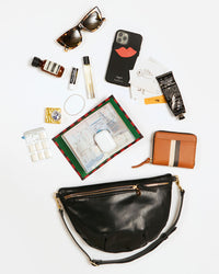 Black Grande Fanny showing the items that it can hold. These items include sunglasses, an aesop sample, chapstick, perfume, gum, an iphone, hand cream, a passport sleeve, airpods and a petit zip wallet