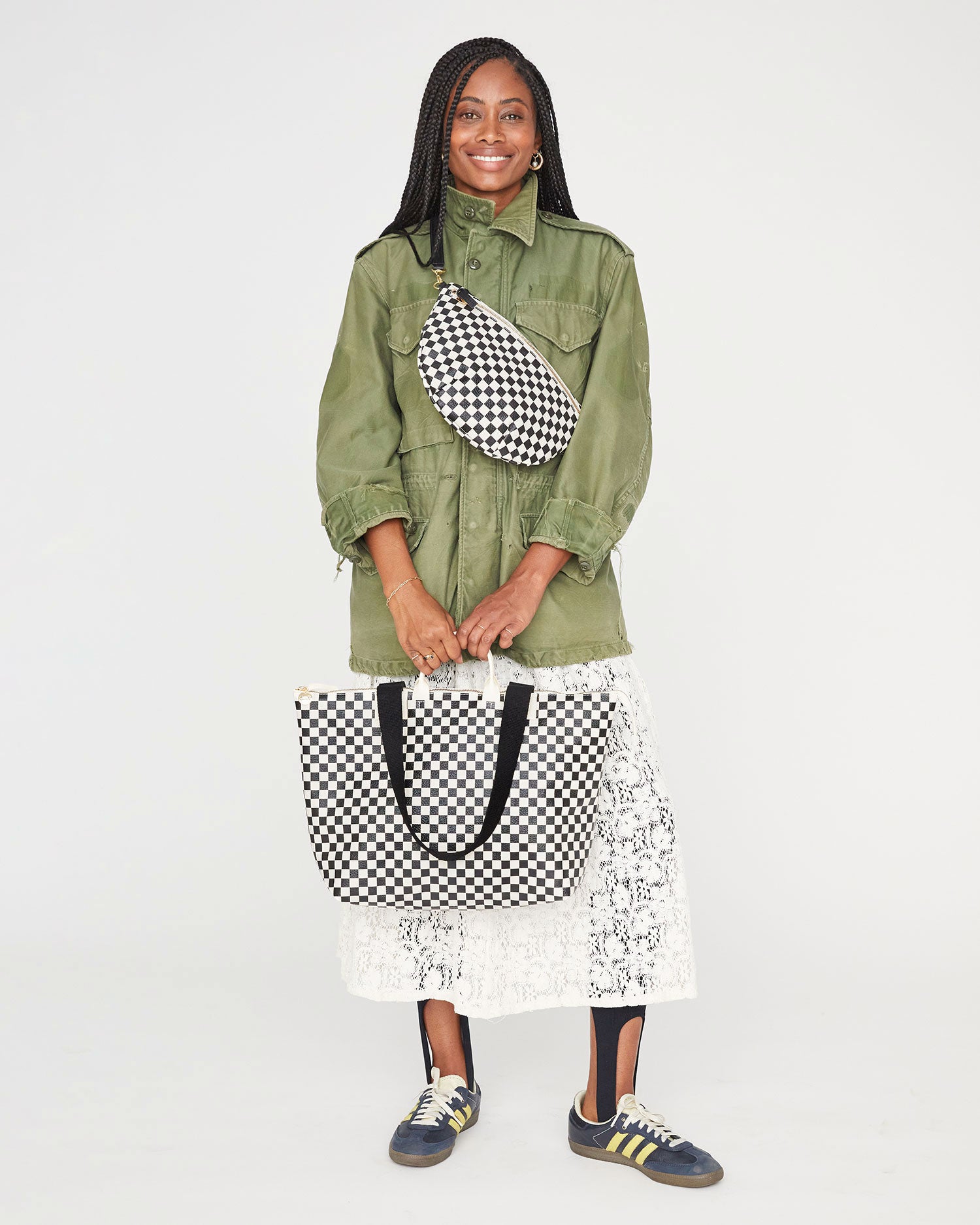 Mecca carrying the Black & Cream Checkers Le Zip Sac by its top handles. she's wearing a cream lace skirt and an army green coat with the black and cream checkers grande fanny worn around her chest